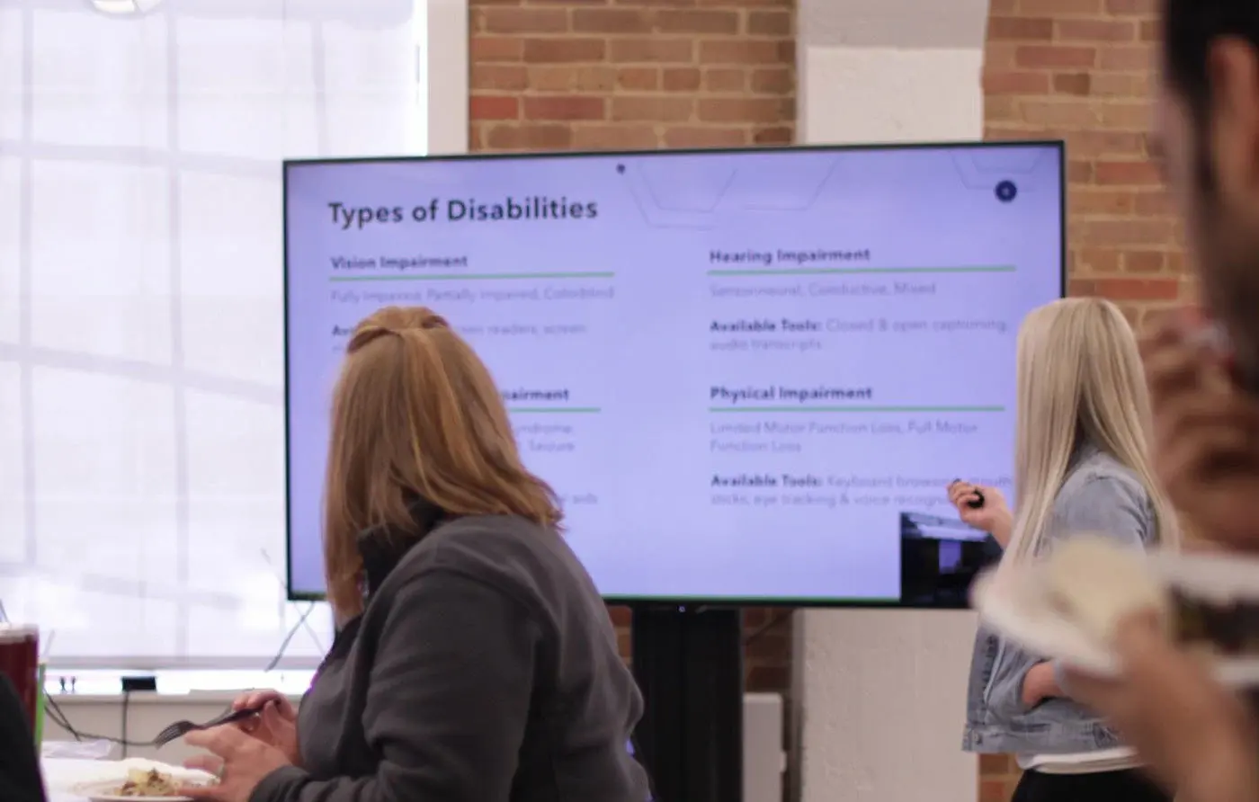 A lunch and learn in a brick-walled office: a screen shows a slide titled Types of Disabilities, with coworkers watching and Kendra presenting beside it.