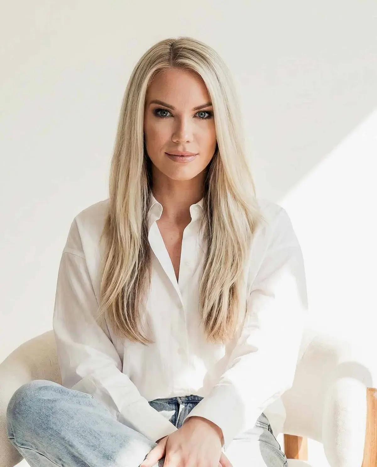 Kendra Shrable, a woman with long blonde hair in a white button-up shirt and jeans, sitting in a cream armchair and looking at the camera.