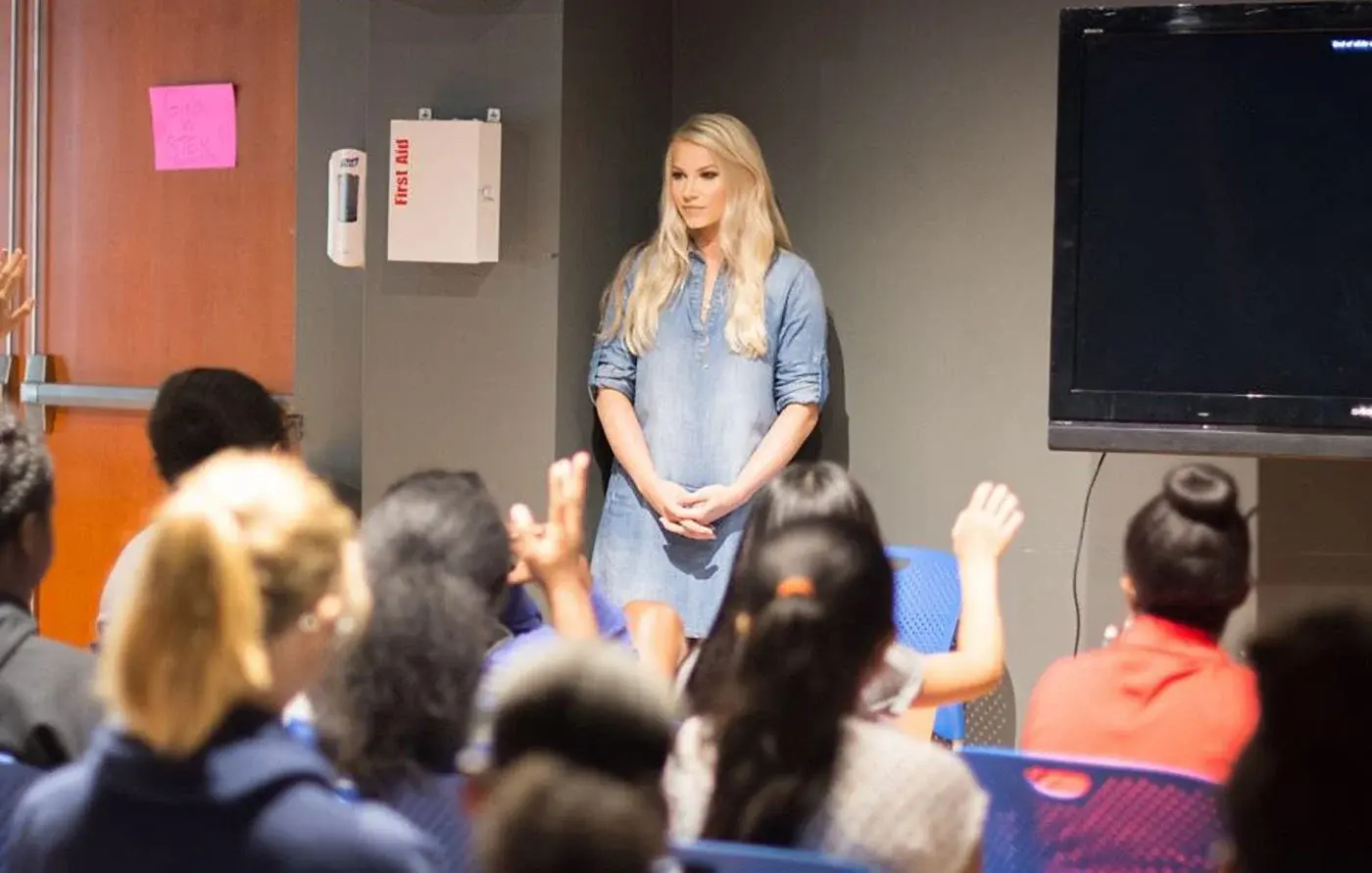 Kendra standing at the front of a room in a denim shirt, speaking to students, several with their hands raised.